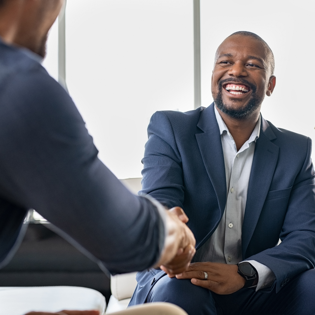 Two professional men shake hands in a bright office, both wearing navy suits and smiling.