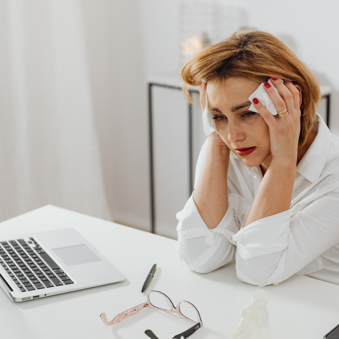 Exhausted woman in a white shirt rests her head in her hands with tissues, at a white desk with a laptop and glasses nearby.