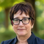 Smiling woman with short brown hair and black-framed glasses, outdoors in a sunny park setting.