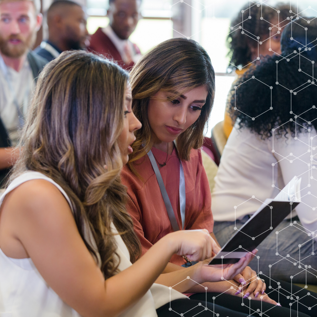 Two women review a tablet together at a conference, discussing notes while others chat in the background.